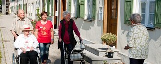 Drei ältere Personen laufen auf dem Gehweg und eine Person wird im Rollstuhl geschoben, begleitet werden sie von einer Mitarbeiterin des Altenheims