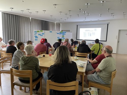 Eine Gruppe Menschen sitzen an zwei Tischen und hören einem Vortrag zu Wahlen zu.