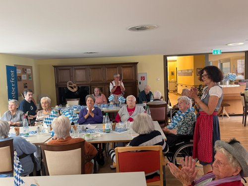 Eine Gruppe älterer Menschen feiert in einem Gemeinschaftsraum ein Oktoberfest. An Tischen mit blau-weißer Dekoration klatschen die Gäste Beifall. Eine Frau im Dirndl applaudiert stehend. Die Atmosphäre wirkt fröhlich und gesellig.
