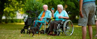 Ein älteres Paar sitzt im Park. Der Mann sitzt auf einem Stuhl, die Frau im Rollstuhl und hält einen blauen Ball. Ein kleiner schwarzer Hund steht neben ihnen. Im Vordergrund steht ein Mann mit dem Rücken zur Kamera.