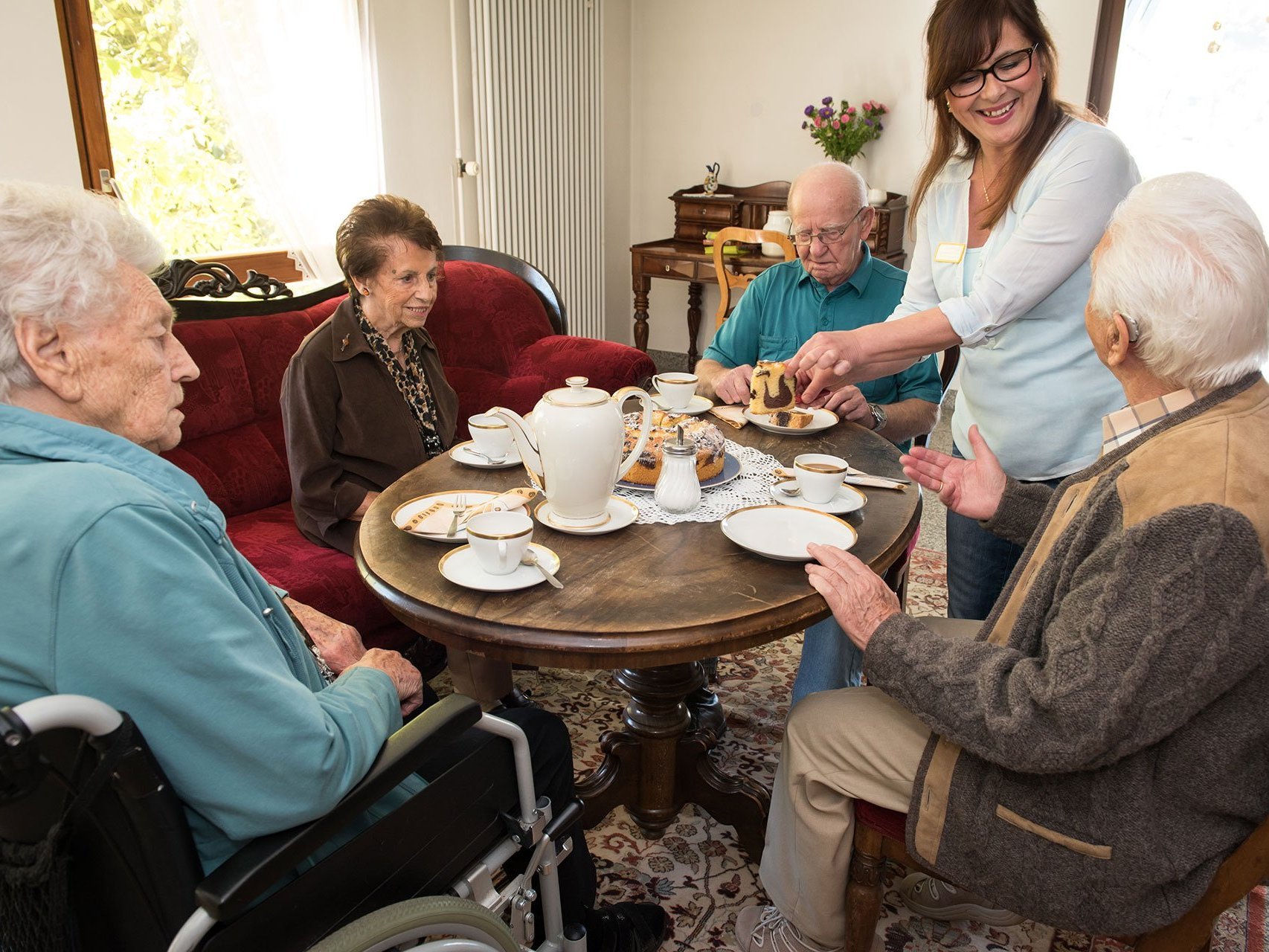 Vier Bewohner sitzen um einen Tisch und halten ein Kaffeekränzchen. Eine Mitarbeiterin des Altenheims schenkt Kaffee ein.