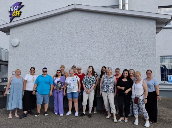 Eine Gruppenaufnahme von etwa 17 Personen vor einem weißen Gebäude mit dem Logo „FACE OFF“. Eine Frau in der Mitte hält eine Urkunde. Die Gruppe besteht aus Männern und Frauen in Freizeitkleidung.