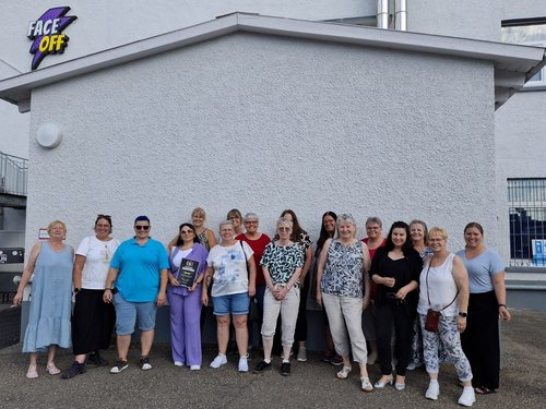 Eine Gruppenaufnahme von etwa 17 Personen vor einem weißen Gebäude mit dem Logo „FACE OFF“. Eine Frau in der Mitte hält eine Urkunde. Die Gruppe besteht aus Männern und Frauen in Freizeitkleidung.