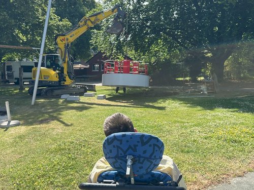 Der Rollstuhlfahrer schaut nun dem Bagger zu wie er das Rollstuhlkarussell in das Erdloch rein hebt