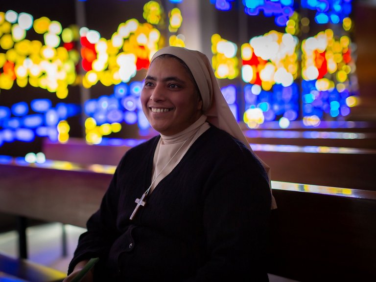 Eine Ordensschwester sitzt auf deiner Kirchenbank und Lächelt. Im Hintergrund schimmern Lichter durch die Kirchfenster.