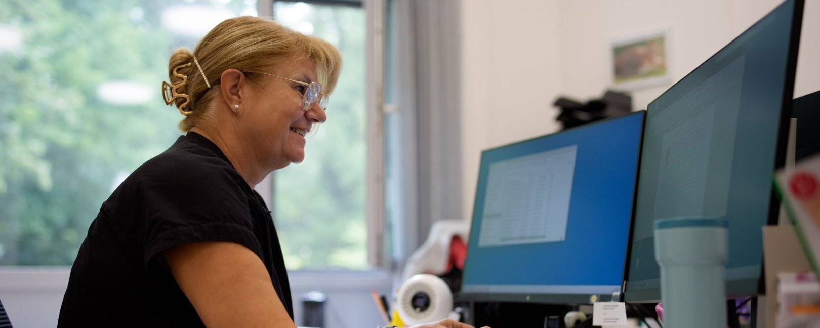 Eine lächelnde Frau mit Brille und blondem Haar sitzt an einem Schreibtisch und arbeitet an zwei Computermonitoren. Sie trägt ein schwarzes Oberteil und tippt auf einer Tastatur. Im Hintergrund ist ein Fenster mit Blick auf Bäume zu sehen.