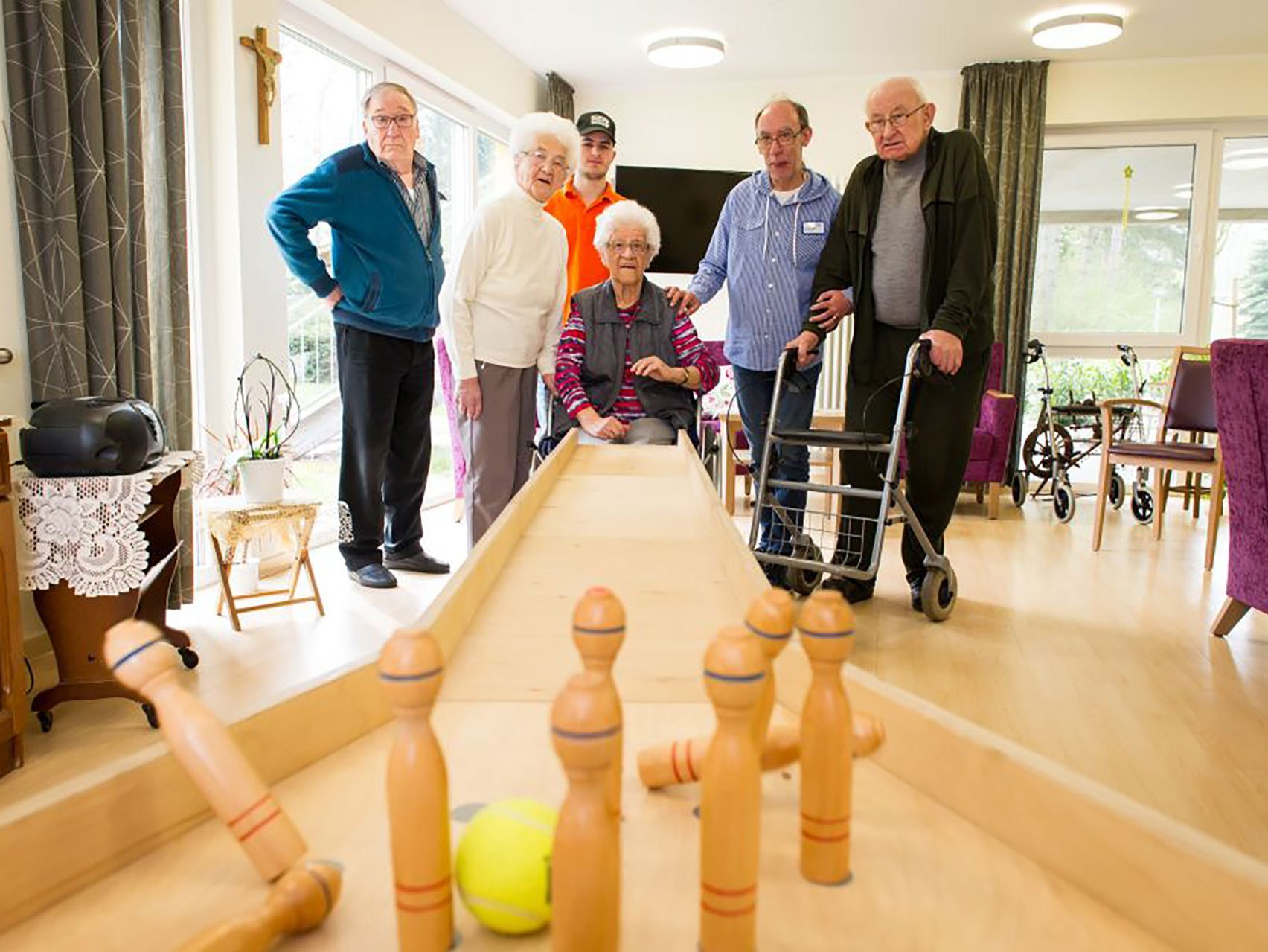 Fünf Bewohner und ein Betreuer stehen um eine mini Kegelbahn herum und Kegel mit einem Tennisball