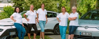 Fünf Frauen in weißen T-Shirts stehen lächelnd vor zwei weißen Fahrzeugen. Auf dem rechten Auto ist der Schriftzug „St. Hildegard“ zu erkennen. Im Hintergrund sind Bäume und ein Gebäude sichtbar.