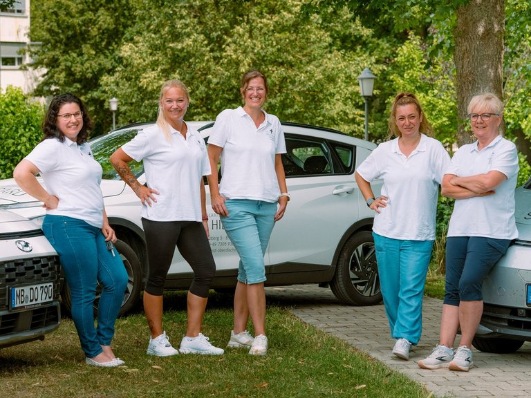 Fünf Frauen in weißen T-Shirts stehen lächelnd vor zwei weißen Fahrzeugen. Auf dem rechten Auto ist der Schriftzug „St. Hildegard“ zu erkennen. Im Hintergrund sind Bäume und ein Gebäude sichtbar.