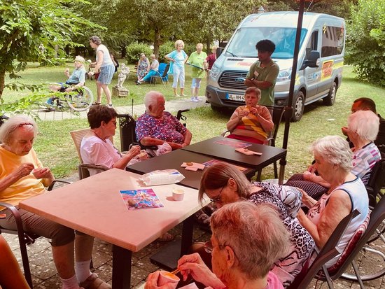 Ältere Menschen sitzen im Freien unter roten Sonnenschirmen an Tischen und essen Eis. Einige nutzen Rollstühle. Im Hintergrund steht ein weißer Van, und weitere Personen entspannen auf einer Wiese.