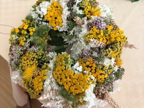 Eine Hand hält einen runden Blumenkranz, der aus leuchtend gelben Blüten, weißen Hortensien und violett-grünen Akzenten besteht. Im Hintergrund sind ein leeres Strohkranzgerüst und weitere grüne Zweige auf einem Tisch zu sehen.
