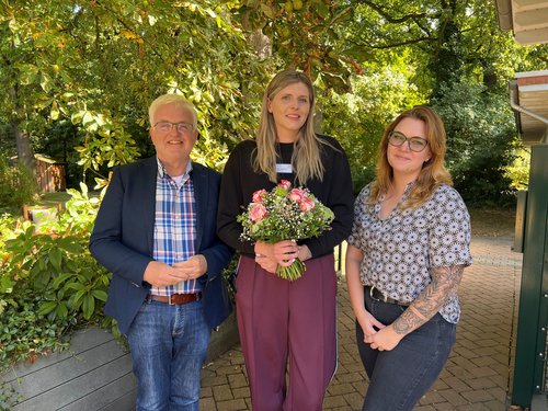 Der Einrichtungsleiter und die zukünftige Einrichtungsleiterin begrüßen eine neue Mitarbeiterin im Team. Sie stehen nebeneinander und die neue Pflegedienstleitung hält einen Blumenstrauß in der Hand.