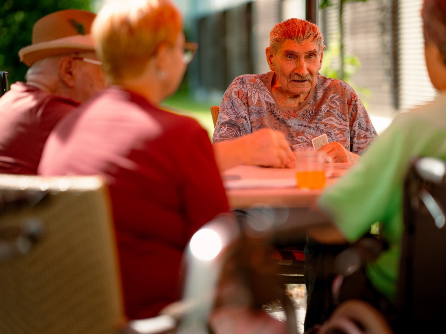 Vier Senioren sitzen im Freien an einem Tisch. Ein älterer Mann mit einem gemusterten Hemd blickt direkt in die Kamera. Die anderen Personen sind von hinten zu sehen. Auf dem Tisch steht ein Glas Orangensaft.