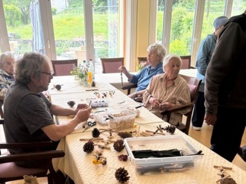 Eine Gruppe älterer Menschen sitzt an einem langen Tisch in einem hellen Raum und bastelt gemeinsam mit Tannenzapfen, Wäscheklammern und anderen Materialien.