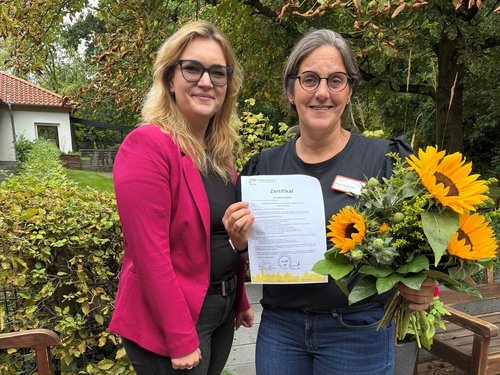 Eine Frau gratuliert einer anderen Frau, die stolz ein Zertifikat in die Kamera hält.