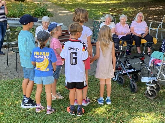 Eine Gruppe von Kindern steht auf einer Wiese und wendet sich einer Gruppe älterer Menschen zu, die in Stühlen sitzen. Die Kinder scheinen eine Aufführung oder Präsentation für die Senioren im Freien zu geben.