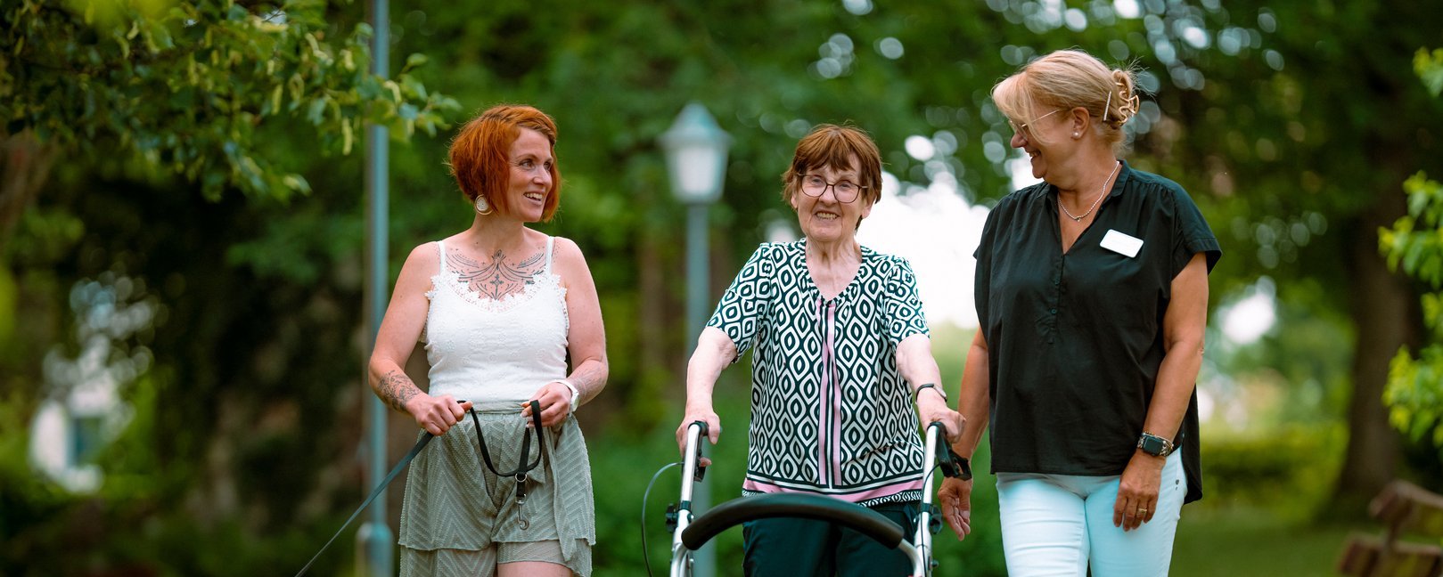 Drei Frauen gehen gemeinsam auf einem Parkweg spazieren. Eine ältere Dame nutzt einen Rollator und wird von zwei jüngeren Frauen begleitet, wobei eine einen kleinen schwarzen Hund führt. Alle lächeln und wirken zufrieden.