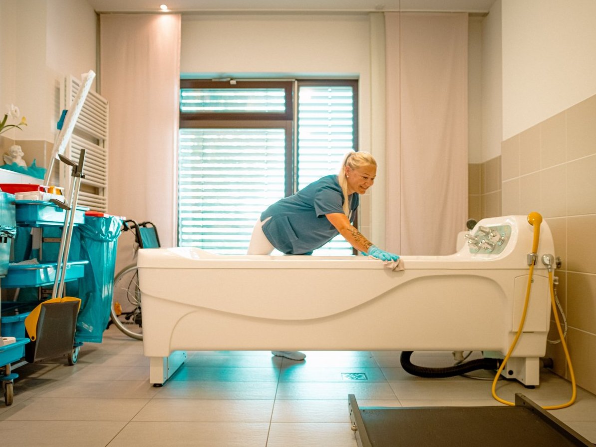 Eine Pflegekraft in blauer Kleidung und Handschuhen reinigt eine große, weiße Spezialbadewanne in einem hellen Pflegezimmer. Im Hintergrund sind ein Rollstuhl und ein Wagen mit Reinigungsmitteln zu erkennen.