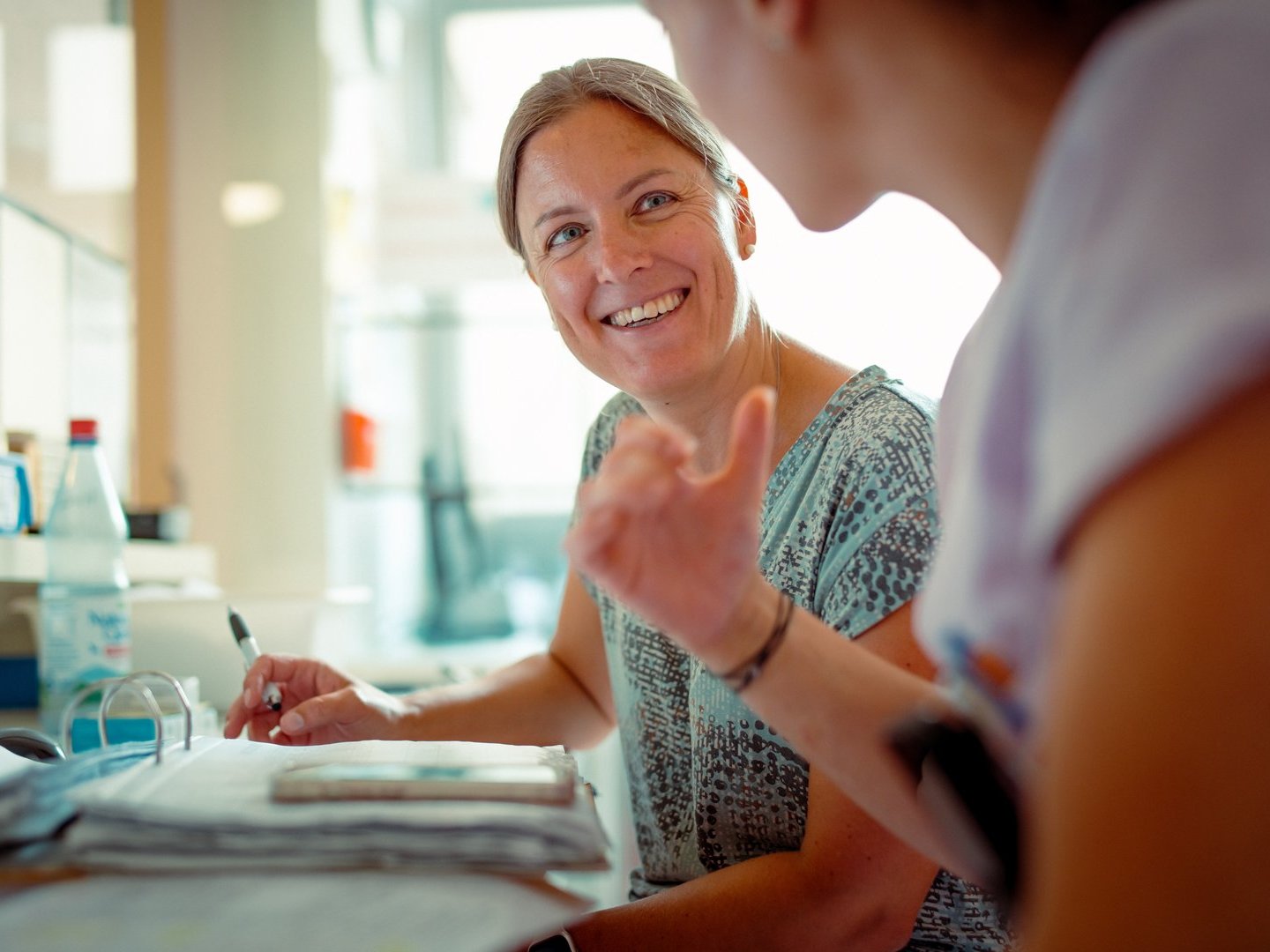 Zwei Frauen führen ein freundliches Gespräch an einem Schreibtisch. Die Frau im Hintergrund lächelt und hält einen Stift, während die Frau im Vordergrund gestikuliert. Die Szene suggeriert eine positive Beratungssituation.