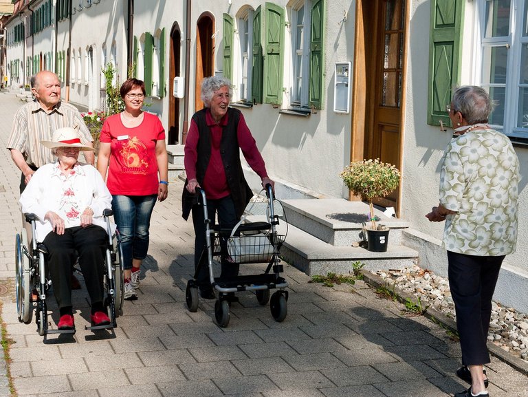 Drei ältere Personen laufen auf dem Gehweg und eine Person wird im Rollstuhl geschoben, begleitet werden sie von einer Mitarbeiterin des Altenheims