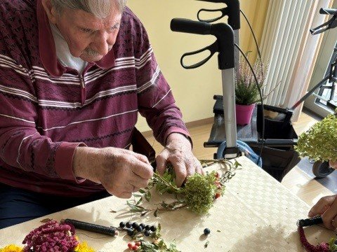 Ein älterer Mann mit weißem Bart und gestreiftem Pullover arrangiert Blumen und Pflanzen an einem Tisch. Vor ihm liegen verschiedene grüne und rote Blüten sowie Beeren. Im Hintergrund ist ein Gehwagen zu sehen.