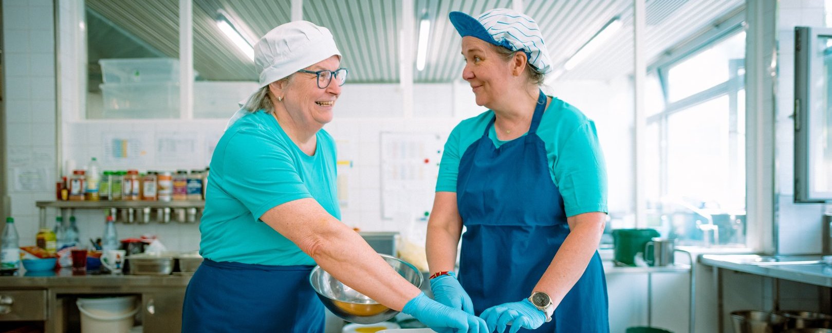 Zwei lächelnde Frauen in blauen Schürzen und Hauben bereiten in einer hellen Großküche gemeinsam Essen vor. Sie stehen an einer Arbeitsplatte mit kleinen Schalen und interagieren freundlich miteinander.