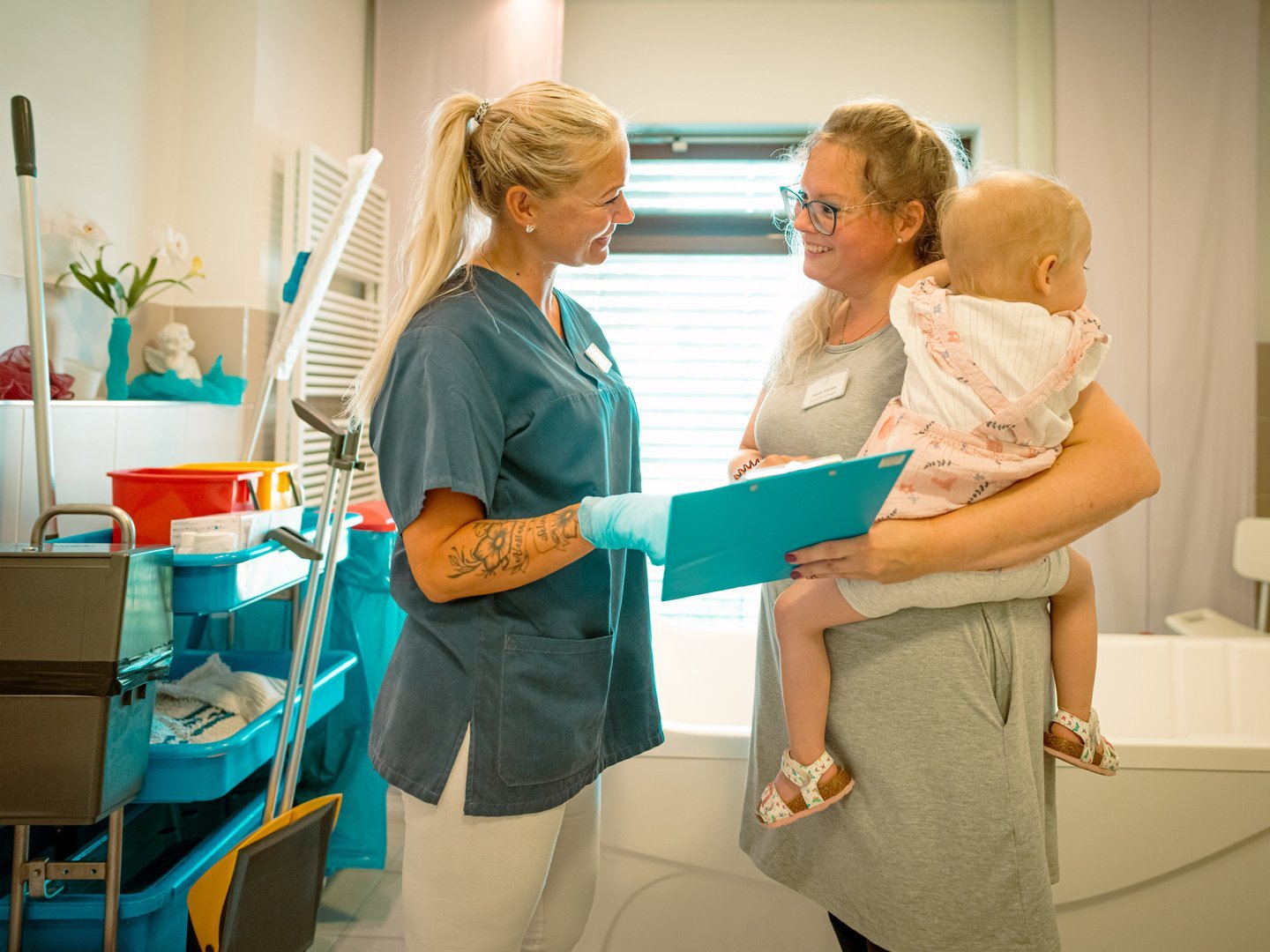 Eine Krankenschwester in blauer Kleidung bespricht lächelnd mit einer Mutter, die ein Baby auf dem Arm hält, Unterlagen auf einer blauen Klemmmappe.