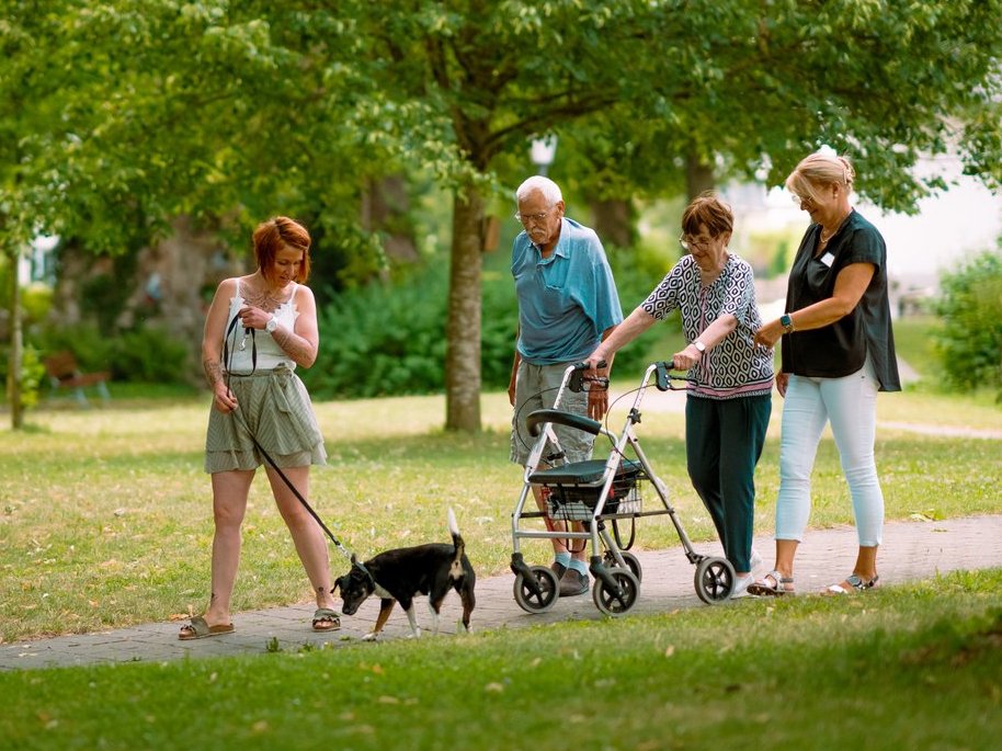 Vier Personen spazieren auf einem Weg durch einen grünen Park. Eine junge Frau führt einen kleinen schwarzen Hund an der Leine. Eine ältere Frau schiebt einen Rollator, begleitet von einer weiteren Frau. Im Hintergrund stehen viele Bäume.
