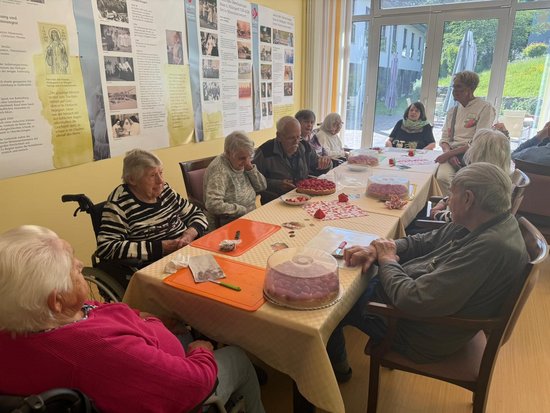 Eine Gruppe älterer Menschen sitzt um einen langen Tisch in einem hellen Raum und genießt verschiedene Kuchen. Es wirkt wie eine gesellige Kaffeestunde in einem Seniorenheim. An der Wand hängen informative Plakate.